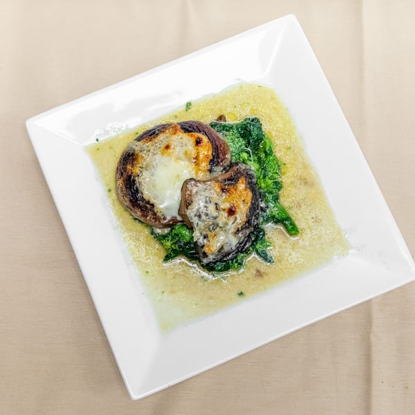 Grilled mushrooms topped with melted cheese, served over leafy greens in a light sauce on a white square plate.