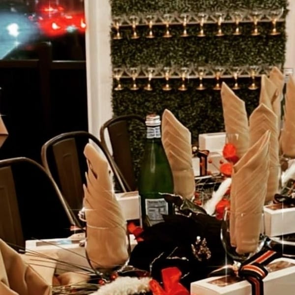 Elegant restaurant dining room with tables set in folded napkins, black plates, gift boxes, flowers, and wine bottles.