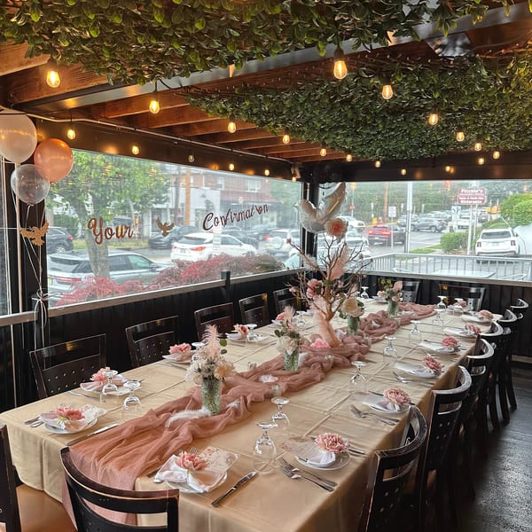 Elegant restaurant dining room with floral table settings, pink linens, greenery-covered ceiling, and glowing pendant lights.