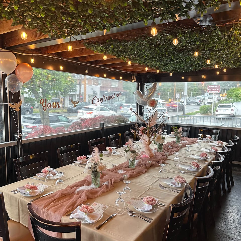 Elegant restaurant dining room with floral table settings, pink linens, greenery-covered ceiling, and glowing pendant lights.