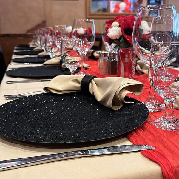 Elegantly set restaurant table with black chargers, beige napkins, glassware, red accents, and floral centerpieces in a warmly lit dining room.