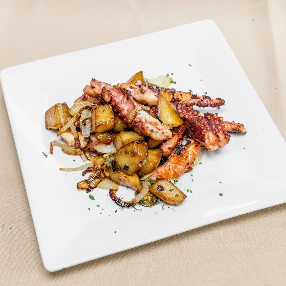Grilled octopus tentacles served with roasted potatoes, onions, and herbs on a white square plate.
