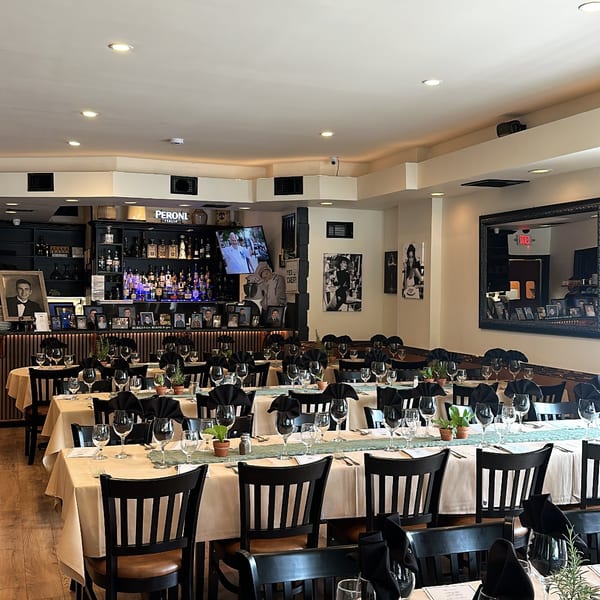 Elegant restaurant dining room with white tablecloths, black napkins, wine glasses, framed photographs, and a fully stocked bar.