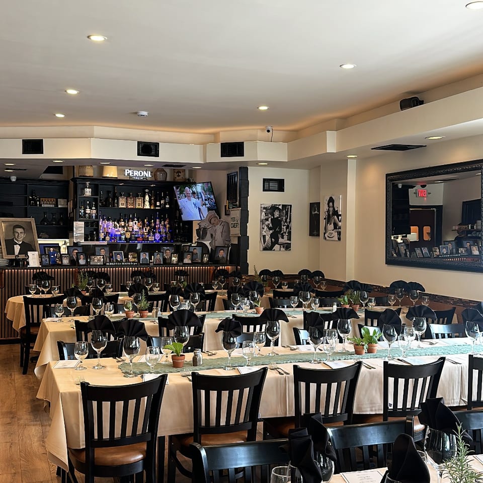 Elegant restaurant dining room with white tablecloths, black napkins, wine glasses, framed photographs, and a fully stocked bar.