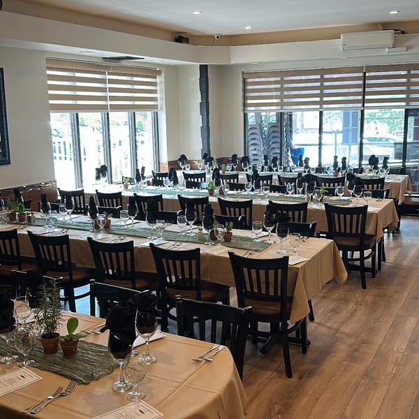 Bright restaurant dining room with neatly set tables, folded black napkins, wine glasses, place settings, and large windows.