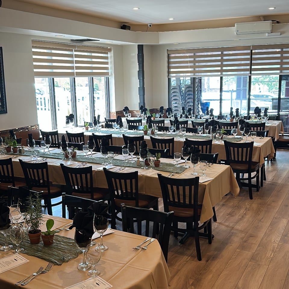 Bright restaurant dining room with neatly set tables, folded black napkins, wine glasses, place settings, and large windows.