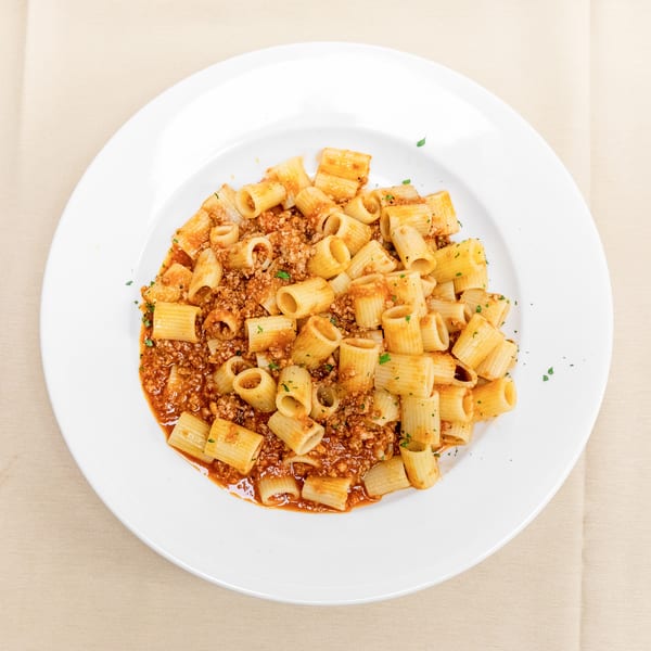 Rigatoni pasta with meat sauce and parsley served on a white plate over a beige tablecloth.