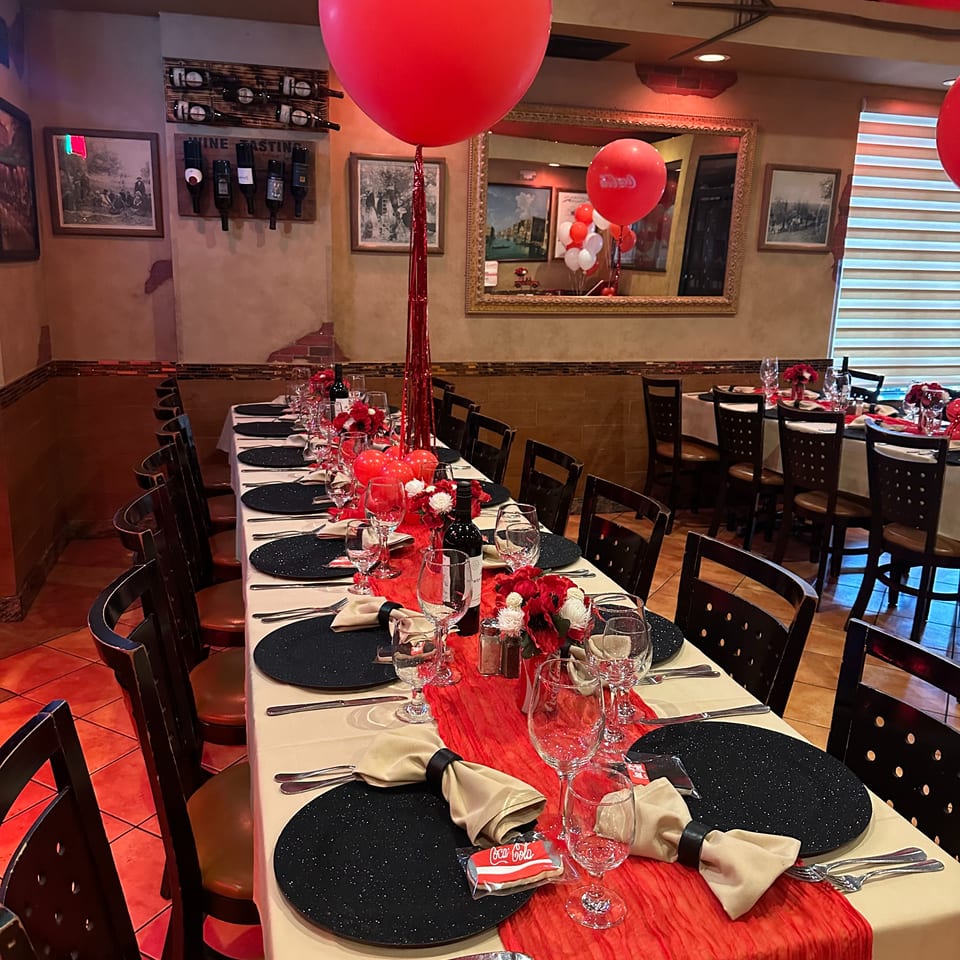 Festively decorated restaurant dining room with red balloons, flower centerpieces, set tables, wine bottles, and warm red lighting.