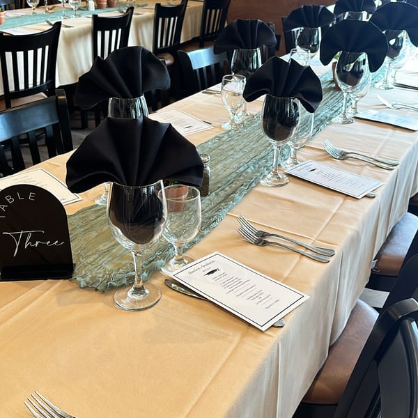 Elegant restaurant dining room with white tablecloths, black napkins, wine glasses, menus, and neatly arranged place settings.