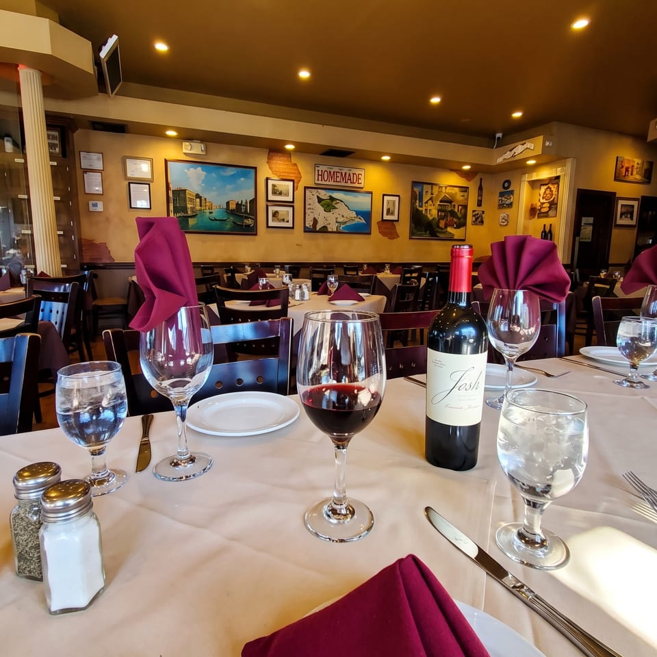 A restaurant table set with wine glasses, a bottle of red wine, and maroon napkins.