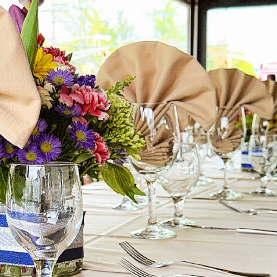 Elegant restaurant dining table set with folded napkins, glassware, silverware, and colorful floral centerpieces by large windows.